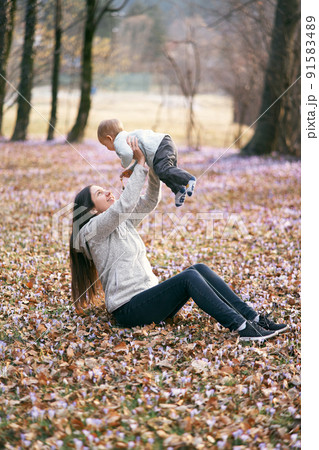 Mom sits on fallen leaves in the park, lifting the baby above her in her arms Mom sits on fallen leaves in the park, lifting the baby above her in her arms 91583489