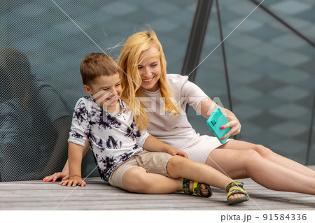 Happy blond woman and little boy sitting on terrace and making selfie on smartphone. Mother and son enjoy time together. Positive young mom spending time with her cute child. Having fun. Family. Happy blond woman and little boy sitting on terrace and making selfie on smartphone. Mother and son enjoy time together. Positive young mom spending time with her cute child. Having fun. Family. 91584336