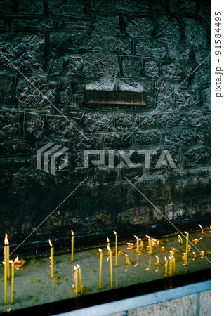 Wax candles burn on a pedestal near a stone wall in a temple 91584495