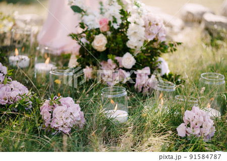 Burning candles in glass jars decorated with flowers stand on green grass Burning candles in glass jars decorated with flowers stand on green grass 91584787