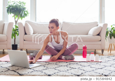 Sporty woman in white and pink sportswear sitting on the floor with dumbbells and a protein shake 91584842