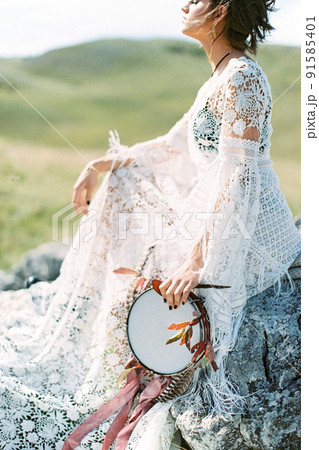 Girl in a dress with a tambourine in her hand sits on stones. Side view 91585401