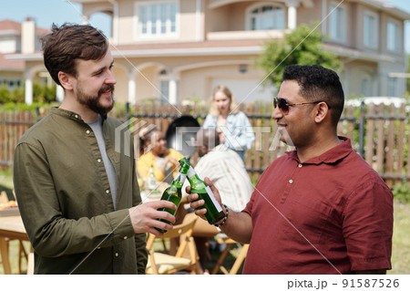 Happy young interracial buddies clinking with bottles of beer at picnic 91587526