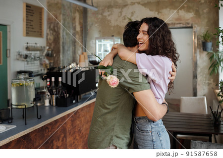 Young affectionate man embracing his cheerful girlfriend with pink rose Young affectionate man embracing his cheerful girlfriend with pink rose 91587658