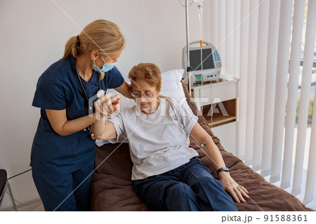Female nurse in mask helping patient to get up from bed in the ward at hospital 91588361