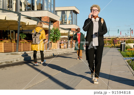 Young woman in eyeglasses walking along the street with beautiful architectural buildings and talking on smartphone Young woman in eyeglasses walking along the street with beautiful architectural buildings and talking on smartphone 91594818