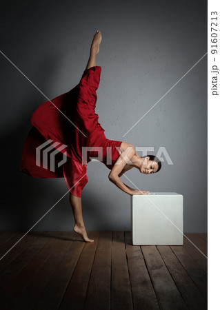 Beautiful expressive woman in red long dress standing splits indoor in studio, grey background, vertical photo, low key. 91607213