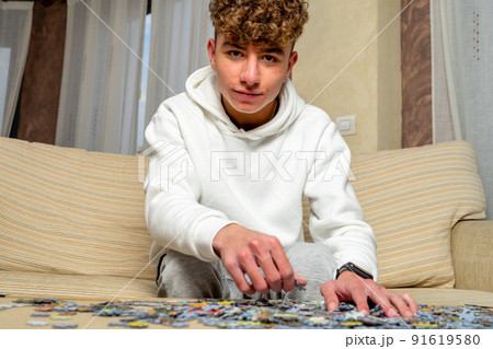 Caucasian young man with curly hair sitting on sofa at home assembling 1000 piece puzzle. Caucasian young man with curly hair sitting on sofa at home assembling 1000 piece puzzle. 91619580