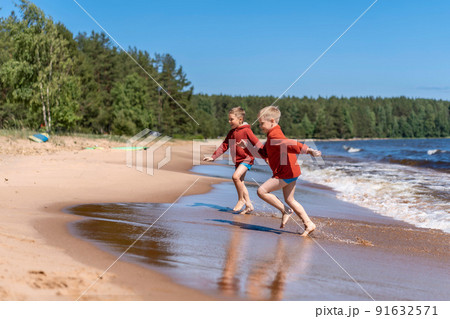 cute caucasian boys wearing red hoodies and blue underpants running from waves in lake with enthusiasm. 91632571