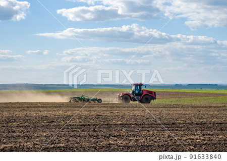 A red farm tractor in a cloud of dust cultivates the soil in the field with a cultivator after harvest. Summer sunny day. Fertile land. Modern agricultural machinery. Copy space. A red farm tractor in a cloud of dust cultivates the soil in the field with a cultivator after harvest. Summer sunny day. Fertile land. Modern agricultural machinery. Copy space. 91633840