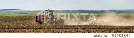 A red farm tractor in a cloud of dust cultivates the soil in the field with a cultivator after harvest. Summer sunny day. Fertile land. Modern agricultural machinery. Copy space. A red farm tractor in a cloud of dust cultivates the soil in the field with a cultivator after harvest. Summer sunny day. Fertile land. Modern agricultural machinery. Copy space. 91633846