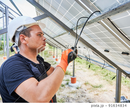 Worker fixing solar panels on metal beams with ropes. Electrician wearing helmet and workwear. Concept of renewable and ecological energy. Modern technology and innovation. Idea of environment safe Worker fixing solar panels on metal beams with ropes. Electrician wearing helmet and workwear. Concept of renewable and ecological energy. Modern technology and innovation. Idea of environment safe 91634834