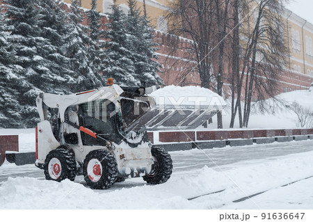 A small loader excavator bobcat removes snow from the sidewalk near the Kremlin walls during a heavy snowfall.  91636647