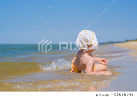 Selective focus on baby girl lying in the water on the sand on the beach. Sunny day. Back view. On the head is a white bandana shawl. Shallow depth of field. Happy childhood. Copy space Selective focus on baby girl lying in the water on the sand on the beach. Sunny day. Back view. On the head is a white bandana shawl. Shallow depth of field. Happy childhood. Copy space 91636648