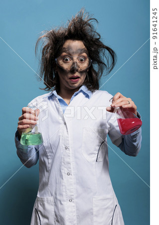 Surprised crazy chemistry specialist holding glass beakers filled with dangerous liquid compounds. Mad chemist with dirty face and messy hair having flasks filled with unknown chemical substances. 91641245