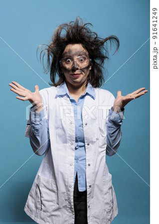 Insane and silly looking crazy scientist with messy hairstyle and dirty face does not understand where experiment went wrong. Mad chemist with goofy expression standing on blue background. Studio shot 91641249