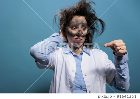 Insane scientist with messy hair and dirty face looking confused at microscope slide tray on blue background. Silly and crazy biochemist looking at microscopic sample after chemical explosion. Insane scientist with messy hair and dirty face looking confused at microscope slide tray on blue background. Silly and crazy biochemist looking at microscopic sample after chemical explosion. 91641251