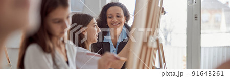 Young beautiful confident teacher is helping a kid to draw on a group lesson in a white modern minimalistic classroom Young beautiful confident teacher is helping a kid to draw on a group lesson in a white modern minimalistic classroom 91643261