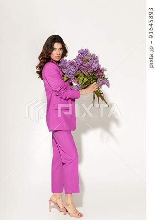 Full-length studio fashion portrait of elegant woman wearing purple suit and holding Lilac brunches against white wall. International womans day, Mothers day and spring concept. Full-length studio fashion portrait of elegant woman wearing purple suit and holding Lilac brunches against white wall. International womans day, Mothers day and spring concept. 91645893