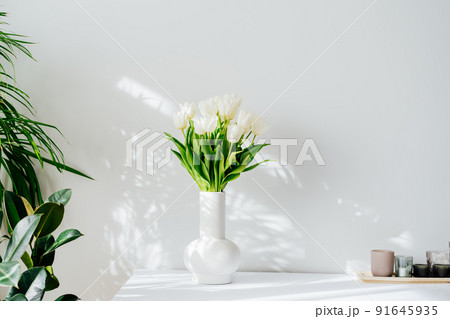 Scandinavian home interior with spring bouquet of white tulip flowers in ceramic vase standing on cabinet. Minimalist design with green plants and white wall with shadows. Biophilia style. Springtime. 91645935