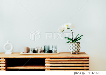 White orchid in blossom and tray with candles on the wooden cabinet against gray wall in modern scandinavian Minimalist home interior. Home decorated with flowers. Selective focus, copy space. White orchid in blossom and tray with candles on the wooden cabinet against gray wall in modern scandinavian Minimalist home interior. Home decorated with flowers. Selective focus, copy space. 91646000