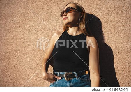 The blonde model wearing sunglasses stands against the beige wall The blonde model wearing sunglasses stands against the beige wall 91648296
