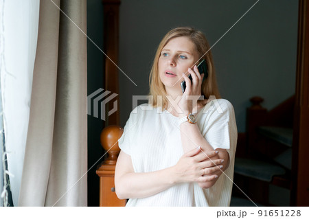 Happy adult woman talking on phone while standing at window against background 91651228