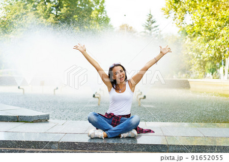 Cheerful young woman in sunglasses sits against backdrop fountain with rainbow 91652055