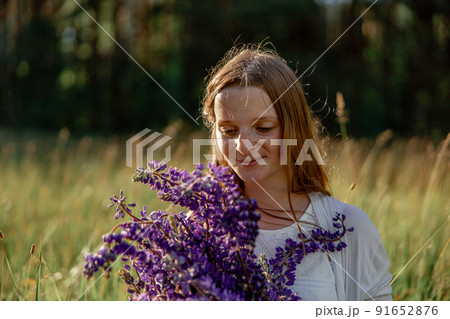Close up portrait of young beautiful redhead woman with freckles, wearing white dress, posing in the nature. Girl with red hair holding flowers. Natural beauty. Diversity, individual uniqueness. 91652876