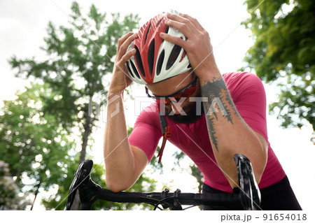 Tired male cyclist in sportswear, glasses and protective helmet walking with his bike in the forest to take a break after riding. Sky blue and forest in the background. 91654018
