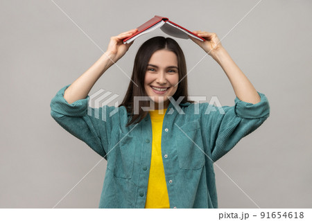 Funny positive woman holding opened book on head, student making ritual to remember lot of study material, wearing casual style jacket. Indoor studio shot isolated on gray background. Funny positive woman holding opened book on head, student making ritual to remember lot of study material, wearing casual style jacket. Indoor studio shot isolated on gray background. 91654618