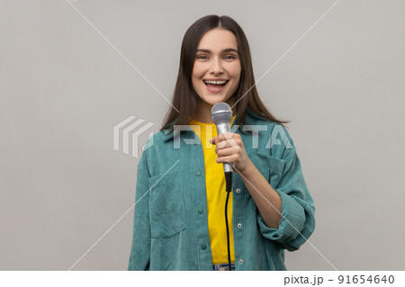 Portrait of positive woman reporter standing with microphone in hands, sing songs with happy expression or telling news, wearing casual style jacket. Indoor studio shot isolated on gray background. 91654640