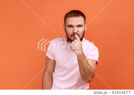 You are liar. Portrait of bearded man pointing finger at nose, showing lie gesture, blaming and suspicion you, body language, wearing pink T-shirt. Indoor studio shot isolated on orange background. You are liar. Portrait of bearded man pointing finger at nose, showing lie gesture, blaming and suspicion you, body language, wearing pink T-shirt. Indoor studio shot isolated on orange background. 91654677