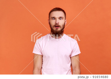 Portrait of bearded man looking cross-eyed with stupid smile, fooling around, making silly face, brainless comical expression, wearing pink T-shirt. Indoor studio shot isolated on orange background. Portrait of bearded man looking cross-eyed with stupid smile, fooling around, making silly face, brainless comical expression, wearing pink T-shirt. Indoor studio shot isolated on orange background. 91654680
