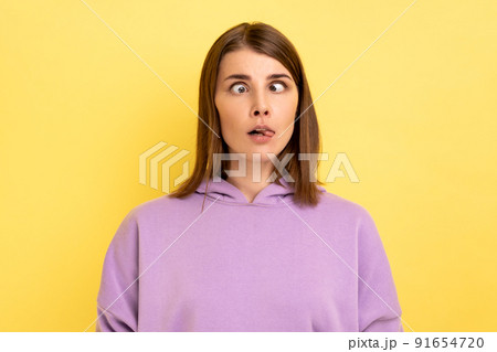 Portrait of young adult foolish woman with hair standing with crossed eyes and tongue out, childish behavior, wearing purple hoodie. Indoor studio shot isolated on yellow background. Portrait of young adult foolish woman with hair standing with crossed eyes and tongue out, childish behavior, wearing purple hoodie. Indoor studio shot isolated on yellow background. 91654720