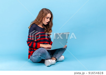 Astonished woman wearing striped casual style sweater, sitting on floor with crossed legs and looking at laptop screen with shocked expression. Indoor studio shot isolated on blue background. 91654828