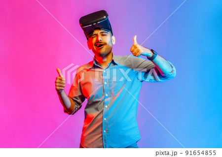 Portrait of man in shirt with virtual reality glasses expressing excitement after playing game simulator, standing with thumbs up. Indoor studio shot isolated on colorful neon light background. Portrait of man in shirt with virtual reality glasses expressing excitement after playing game simulator, standing with thumbs up. Indoor studio shot isolated on colorful neon light background. 91654855