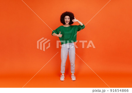 Full length portrait of happy friendly woman with Afro hairstyle wearing green casual style sweater standing with thumbs up, expressing positive. Indoor studio shot isolated on orange background. Full length portrait of happy friendly woman with Afro hairstyle wearing green casual style sweater standing with thumbs up, expressing positive. Indoor studio shot isolated on orange background. 91654866