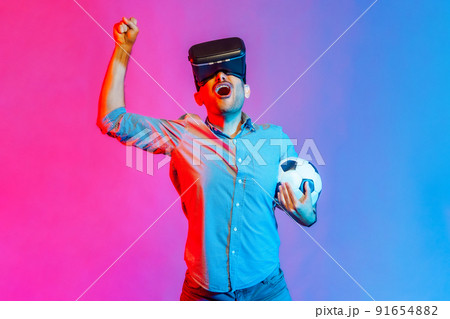 Portrait of man in shirt wearing headset and holding soccer ball, watching 3d football team, cheering, celebrating victory. Indoor studio shot isolated on colorful neon light background. 91654882