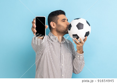 Portrait of bearded handsome man standing kissing soccer ball and showing mobile phone with empty black display, wearing striped shirt. Indoor studio shot isolated on blue background. 91654945