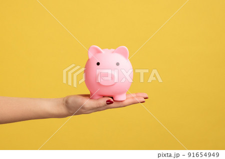 Closeup profile shot of woman hand holding pink piggy bank, investment, saving money, currency, deposit. Indoor studio shot isolated on yellow background. Closeup profile shot of woman hand holding pink piggy bank, investment, saving money, currency, deposit. Indoor studio shot isolated on yellow background. 91654949