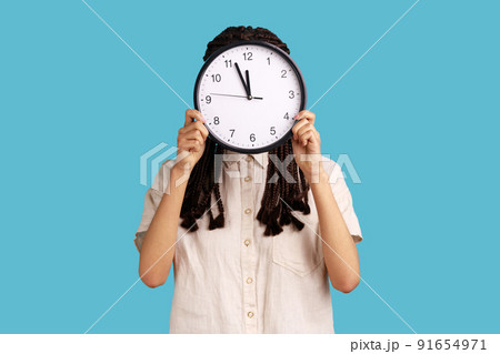 Time to work. Woman with dreadlocks hiding face behind big wall clock, time management, schedule and business meeting appointment, wearing white shirt. Indoor studio shot isolated on blue background. 91654971