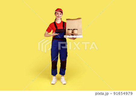 Full length portrait of courier woman presenting coffee and boxes with pizza, offering drinks and food delivery, wearing overalls and red cap. Indoor studio shot isolated on yellow background. Full length portrait of courier woman presenting coffee and boxes with pizza, offering drinks and food delivery, wearing overalls and red cap. Indoor studio shot isolated on yellow background. 91654979
