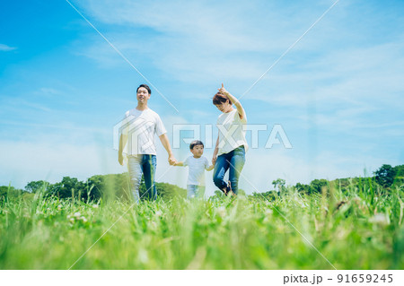 手を繋いで晴天の緑地を歩く親子3人 91659245