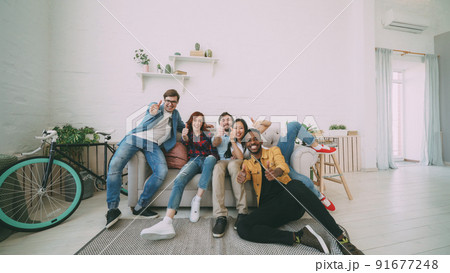 Young friends sitting on sofa smiling and showing thumbs up all together at home indoors 91677248