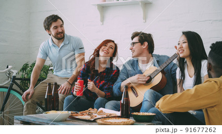 Happy young friends have party at shared apartment and singing together while their friend playing guitar at home indoors 91677262
