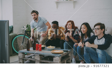 Group of young friends watching olympic games match on TV together eating snacks and drinking beer on holidays at home indoors. Some of them happy with their team winning but others disappointed 91677321