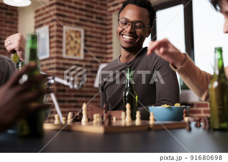 Smiling heartily man sitting at table in living room while enjoying chess game victory. Smart looking person sitting at home with multiracial competitive friends while relaxing with leisure activity. Smiling heartily man sitting at table in living room while enjoying chess game victory. Smart looking person sitting at home with multiracial competitive friends while relaxing with leisure activity. 91680698