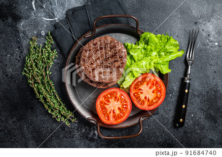 Tasty grilled burger beef patty with tomato, spices and lettuce in kitchen tray. Black background. Top view 91684740
