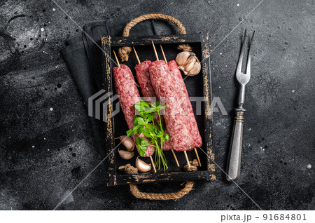 Fresh raw beef meat kebabs sausages on skewers in wooden tray. Black background. Top view Fresh raw beef meat kebabs sausages on skewers in wooden tray. Black background. Top view 91684801